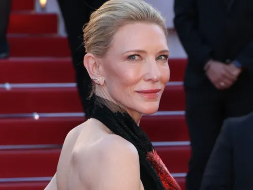 Cate Blanchett looks over her shoulder to smile at the camera. She is wearing a black evening gown with an open back, fringe sleeves, and dramatic floral embroidery in multiple colors. Her blonde her is pulled back into an elegant updo.
