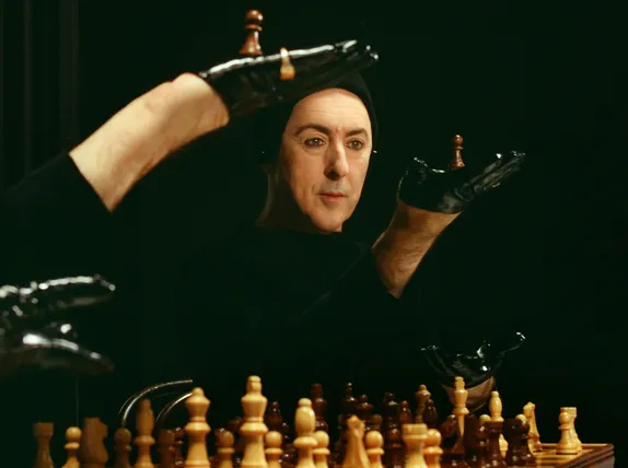 Alan Cumming playing chess mimelike gestures with gloved hands