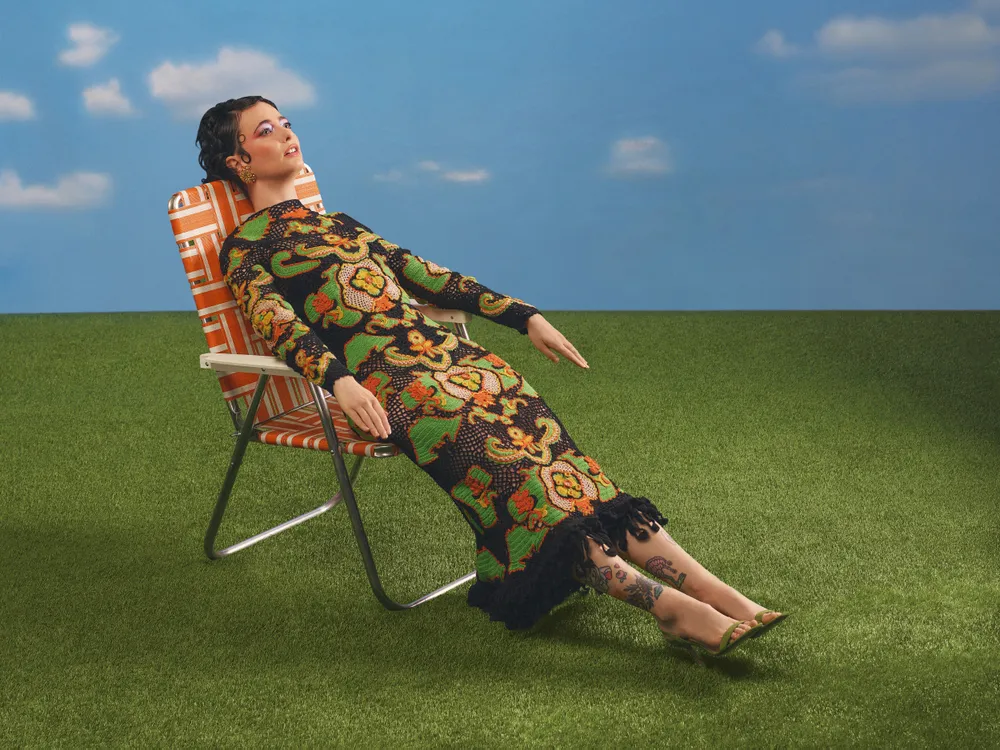 Sarah Sherman in a floralpatterned dress reclining on a lawn chair on artificial grass backdrop of a blue sky with clouds
