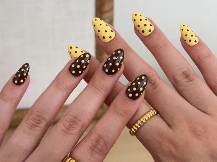 Hands with manicured nails displaying alternating polkadot patterns on yellow and dark bases adorned with rings
